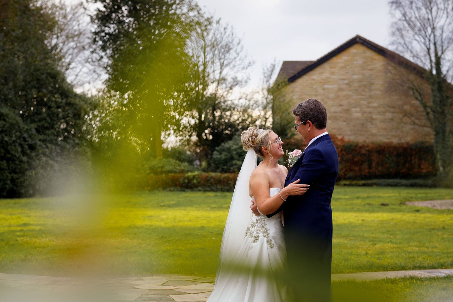 A romantic wedding couple embraces on a lush green lawn with a stone building in the background.