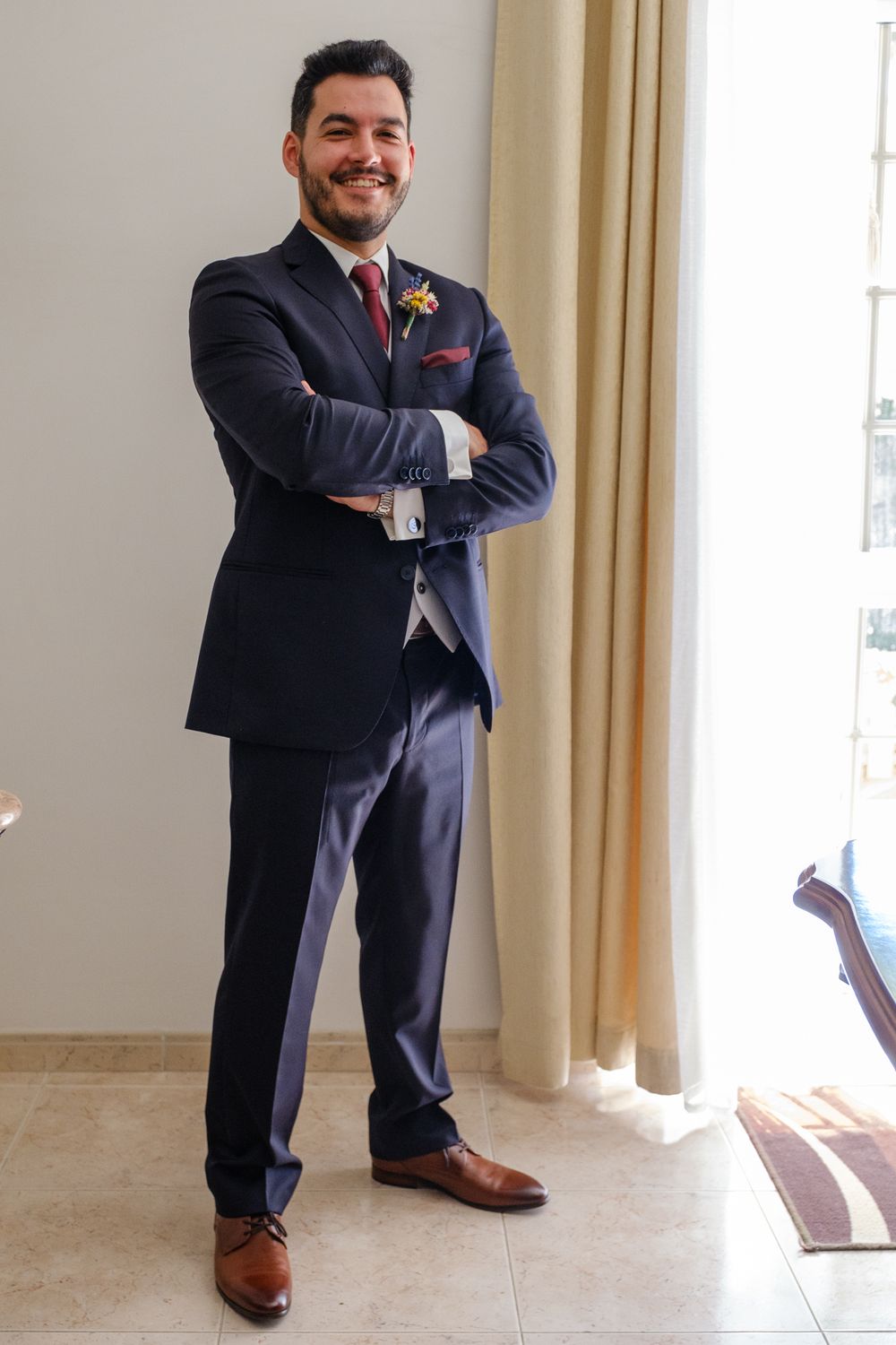 A person in a navy suit with brown shoes stands confidently with arms crossed.