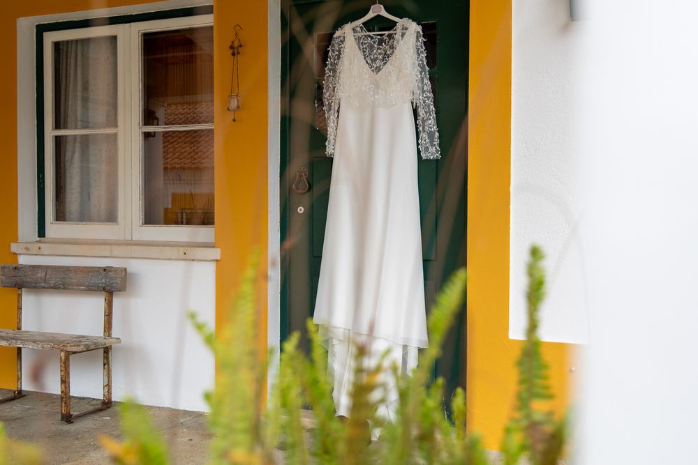 A white lace wedding dress hangs elegantly against a yellow wall with a window frame and greenery in view.