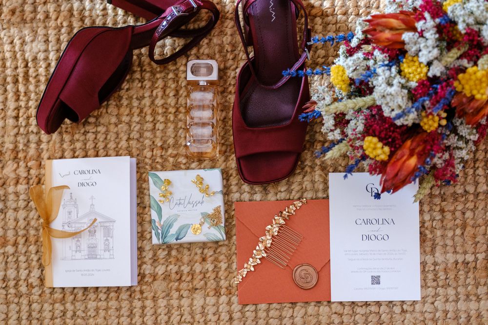 Wedding accessories including burgundy shoes, invitations and a colorful dried flower bouquet arranged on woven material.