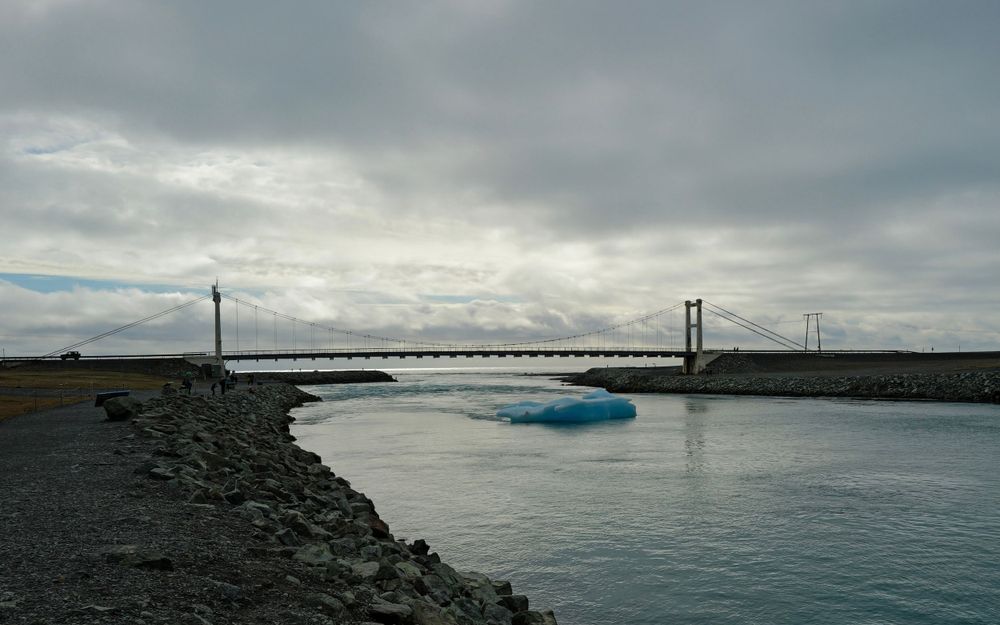Bridge spans across water with floating ice under cloudy skies in Iceland.