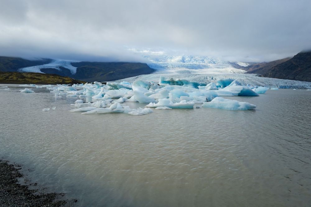 Massive blue icebergs float in a glacial lake surrounded by mountains under cloudy skies.
