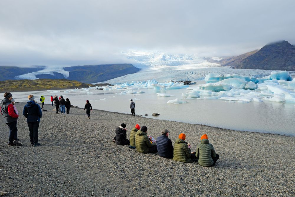 Tourists gather along a rocky shore to view massive blue icebergs floating in a glacial lagoon beneath cloudy mountains.