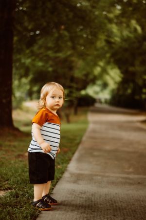A small child stands on a pathway wearing a striped shirt with orange sleeves and dark shorts.