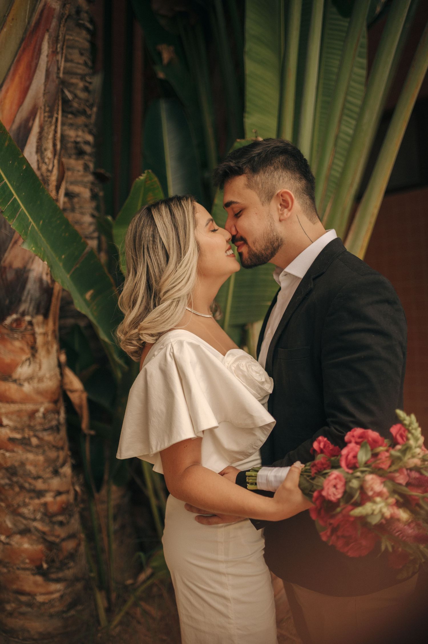 Romantic couple in formal wear embraces near tropical plants and flowers.