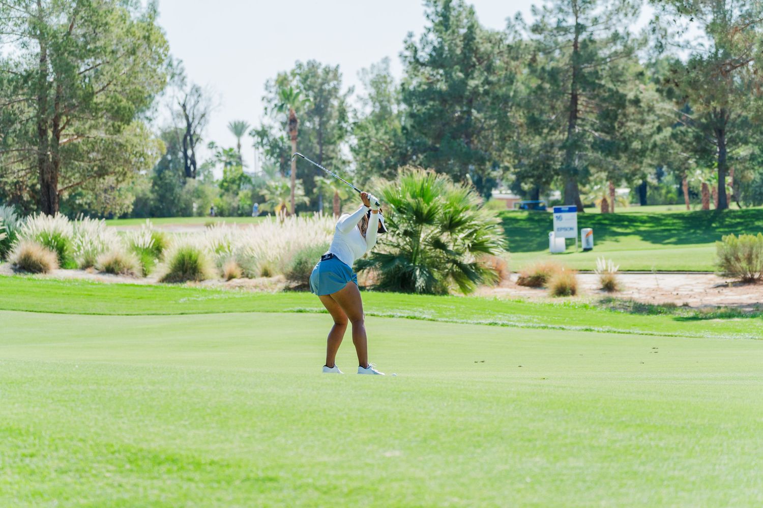 A golfer takes a swing on a lush green fairway surrounded by palm trees and ornamental grasses on a sunny day.