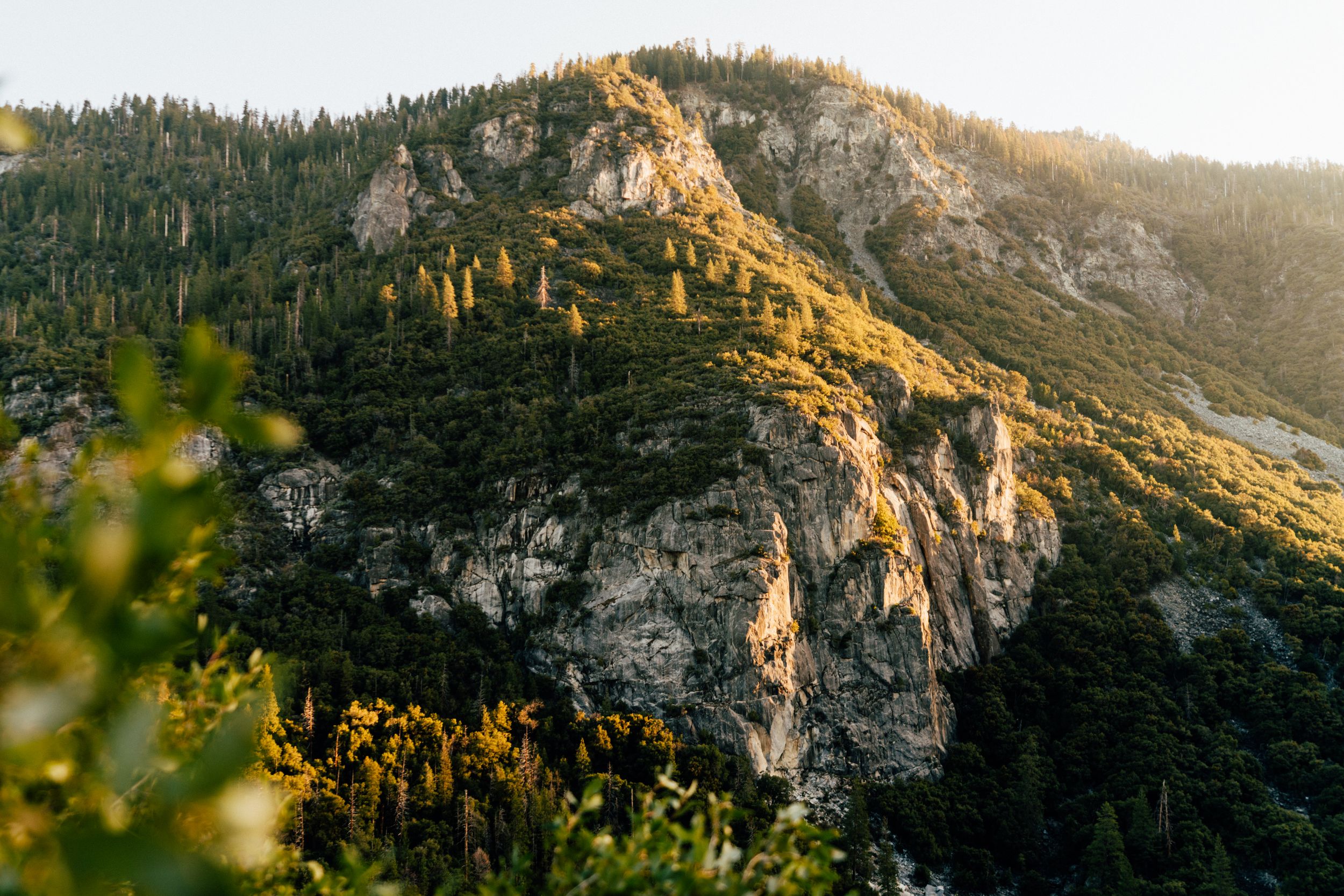 Dramatic mountain landscape at sunset with golden light illuminating rocky peaks and forested slopes.