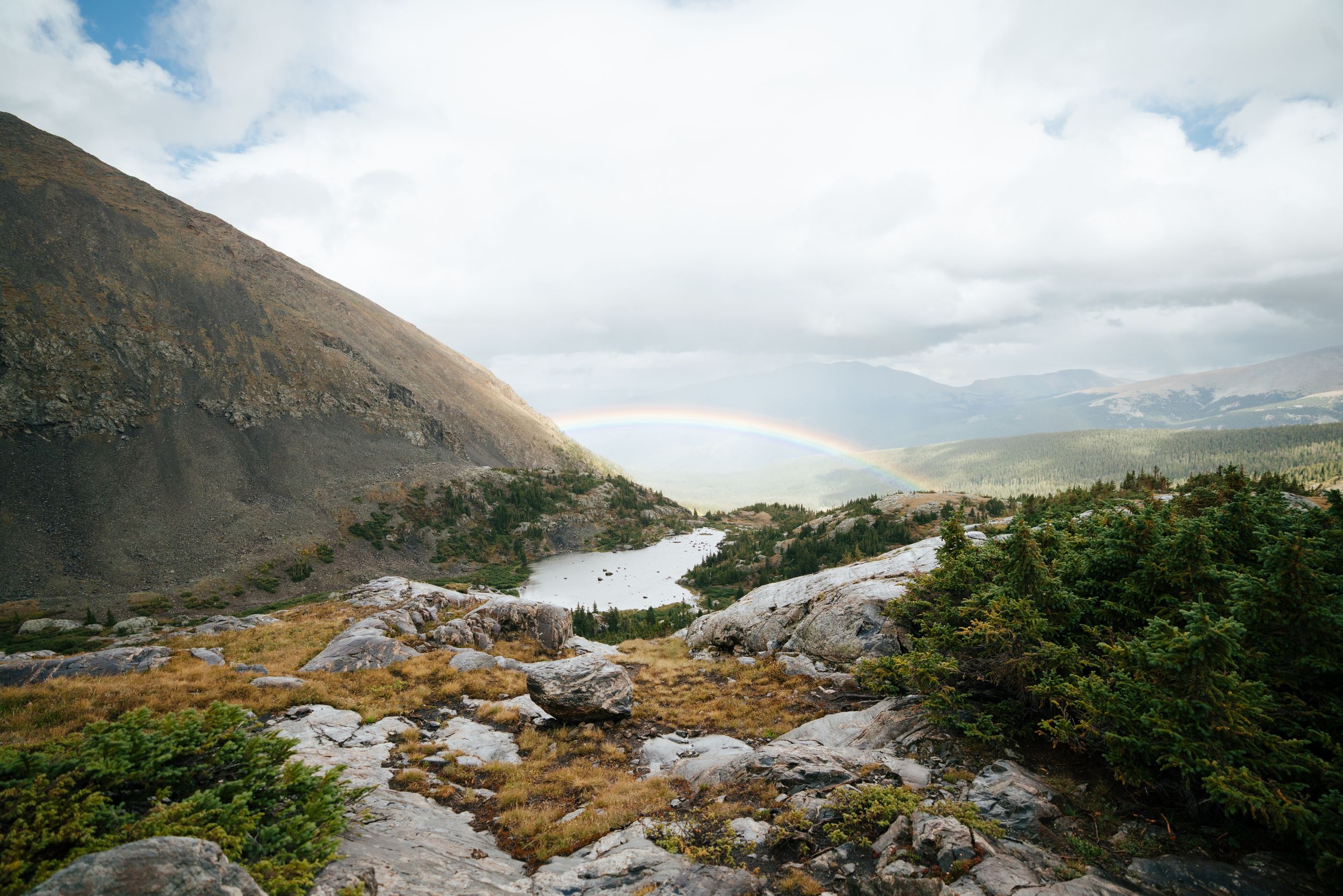 Panoramic mountain landscape with rocky terrain and winding river flowing through rugged wilderness under cloudy skies.