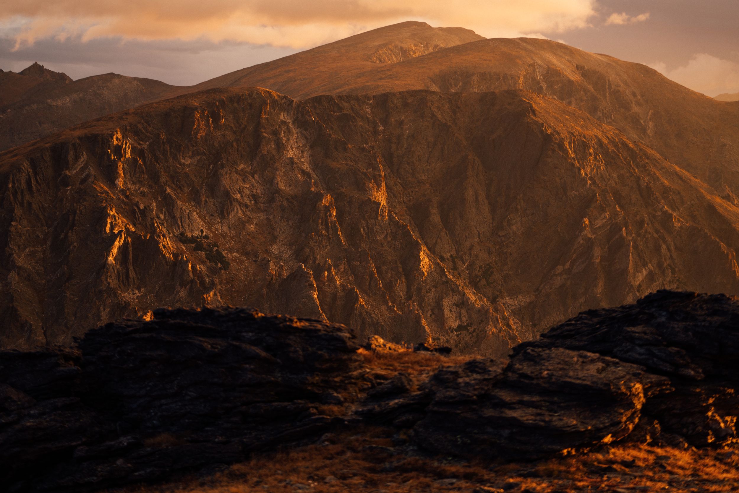 Majestic mountain range bathed in warm sunset light with dramatic cloud formations overhead.