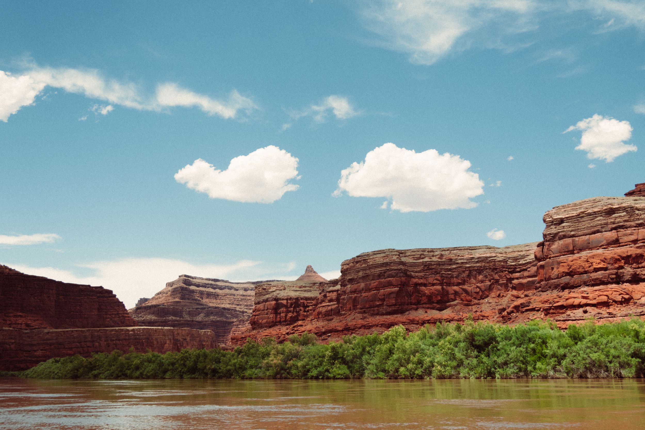 Red rock formations and green vegetation line a river in desert landscape.