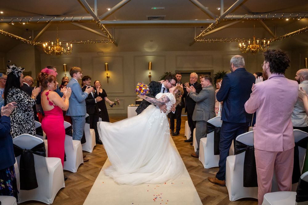 Bride and groom walk down the aisle as wedding guests applaud in an elegant chandelier-lit reception hall.