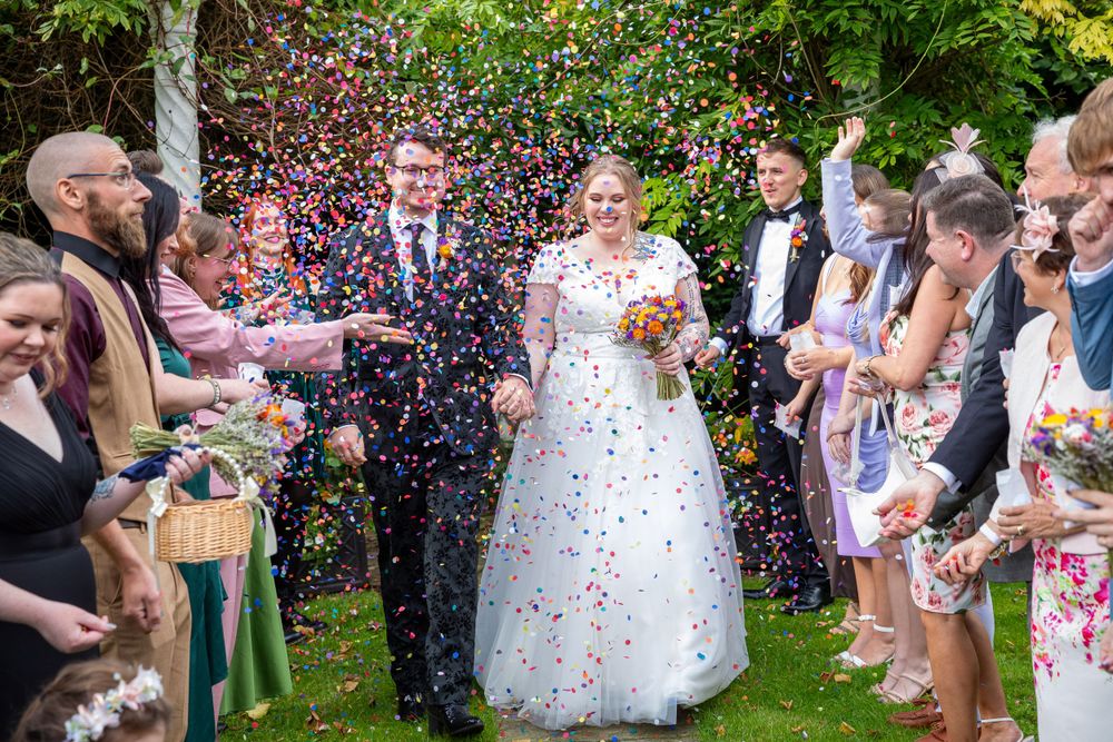 Joyful bride and groom showered with colorful confetti by guests in a sunny outdoor garden wedding.