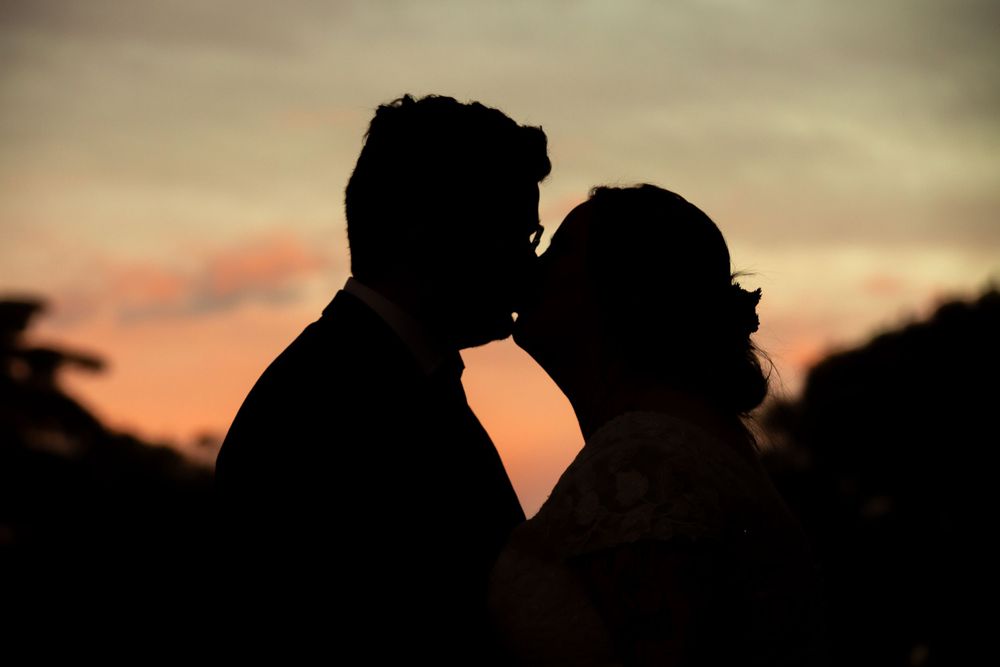 Romantic silhouette of bride and groom sharing a kiss against a stunning sunset sky.