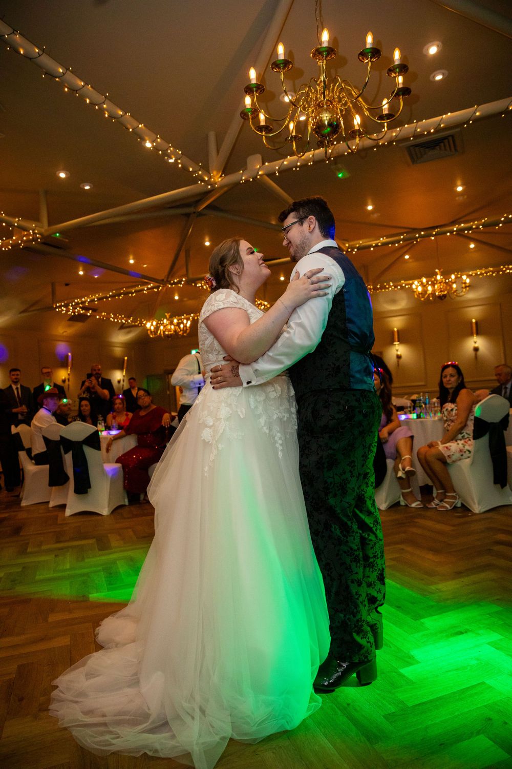 Bride and groom share their first dance under fairy lights and a chandelier in a warmly lit wedding venue.