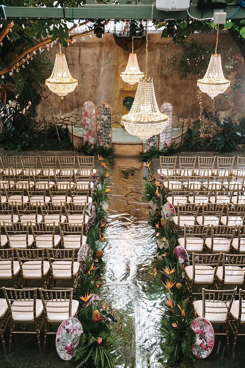 Elegant wedding ceremony setup with crystal chandeliers and mirrored aisle lined with wooden chairs.