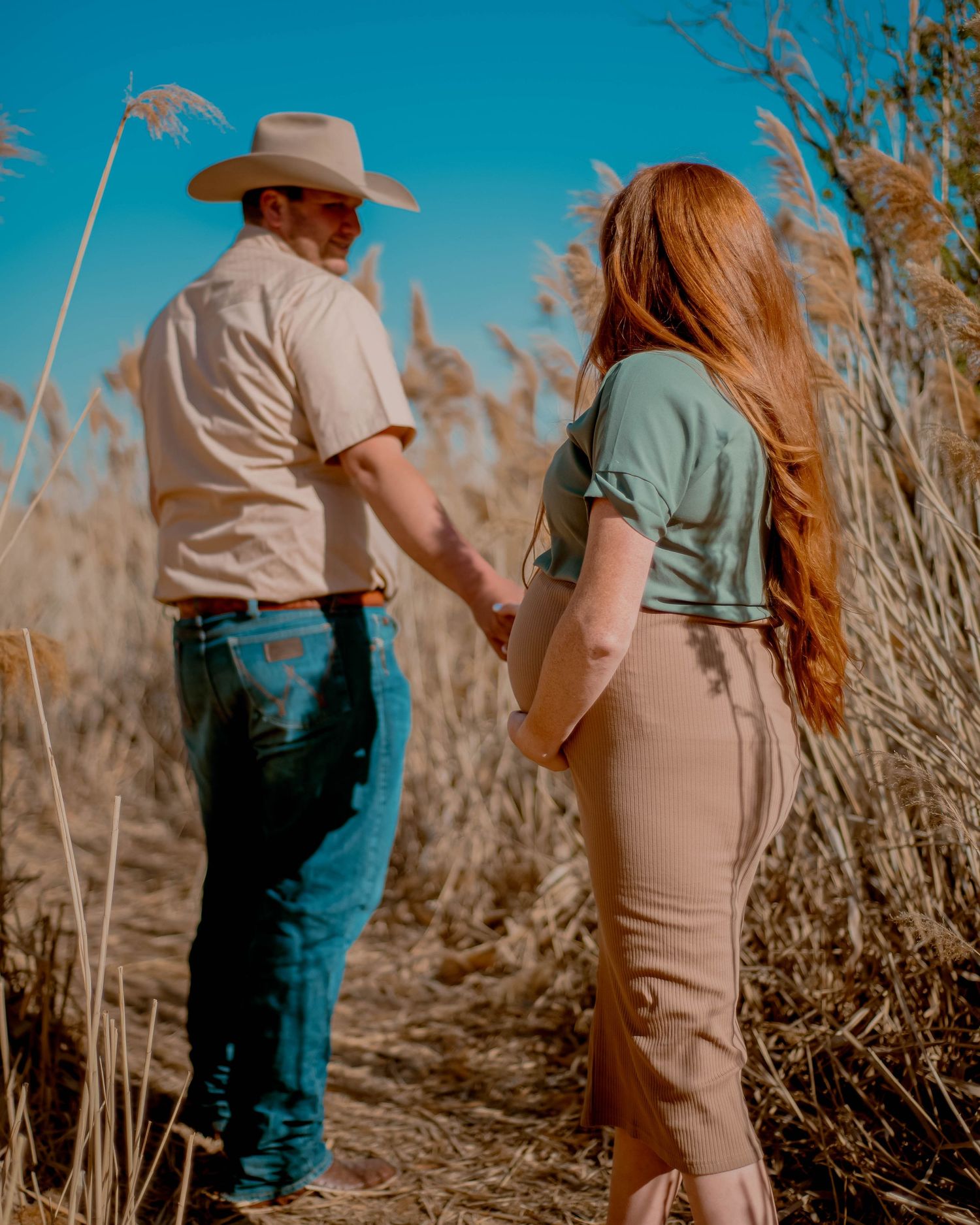 Couple in casual western wear walking through golden grass field on sunny afternoon.
