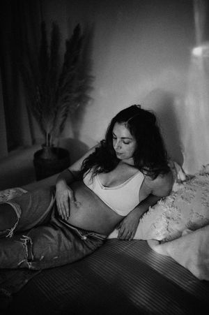 Black and white photograph of a person reclining on a bed in casual attire, with a houseplant visible in the background.