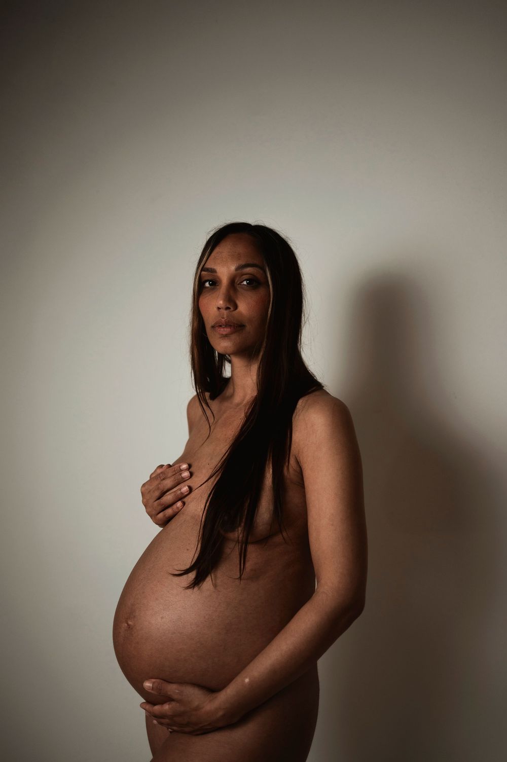 Artistic maternity portrait in moody lighting captures an intimate silhouette against a neutral wall.