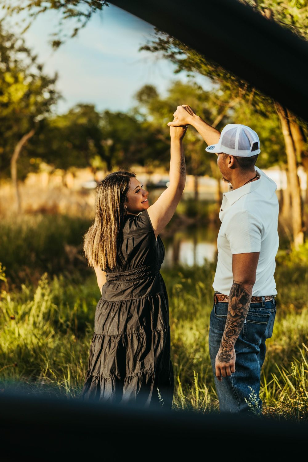 A couple dances outdoors at golden hour, man in white tee and cap, woman in black dress, trees and pond behind them.