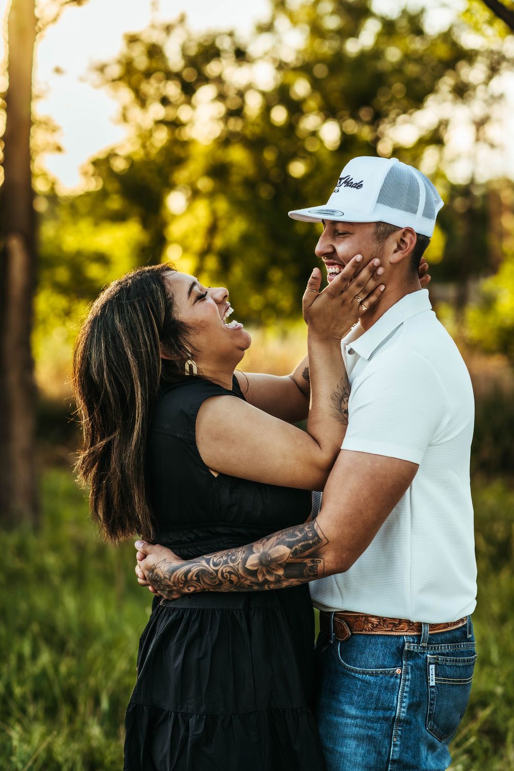 Couple laughing and embracing outdoors at golden hour, woman with tattoos holding man's face, both smiling joyfully.