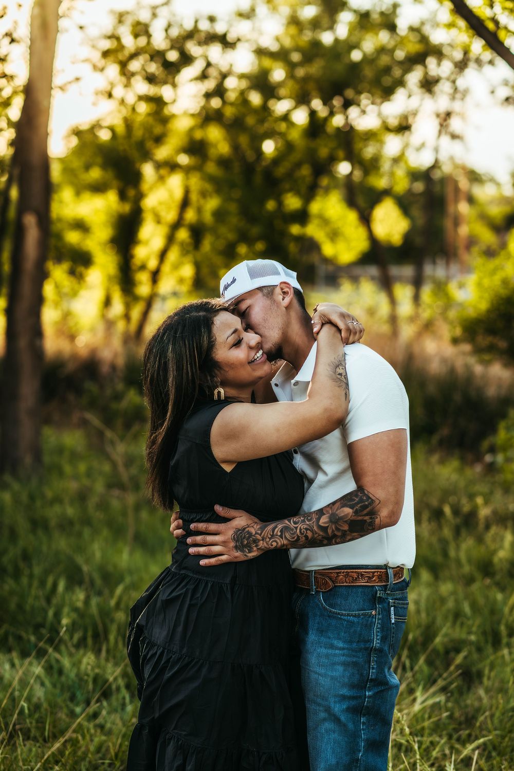 Couple embracing outdoors at golden hour, man with tattoos in white shirt and jeans, woman in black dress, green trees background.