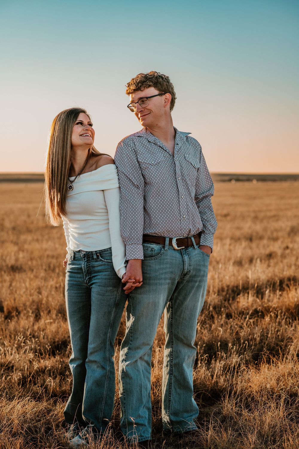 A couple holds hands while standing in a golden wheat field at sunset against a gradient blue and pink sky.