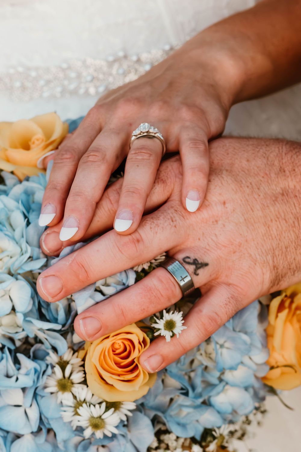 Wedding rings and hands rest on a bouquet of blue hydrangeas and peach roses.