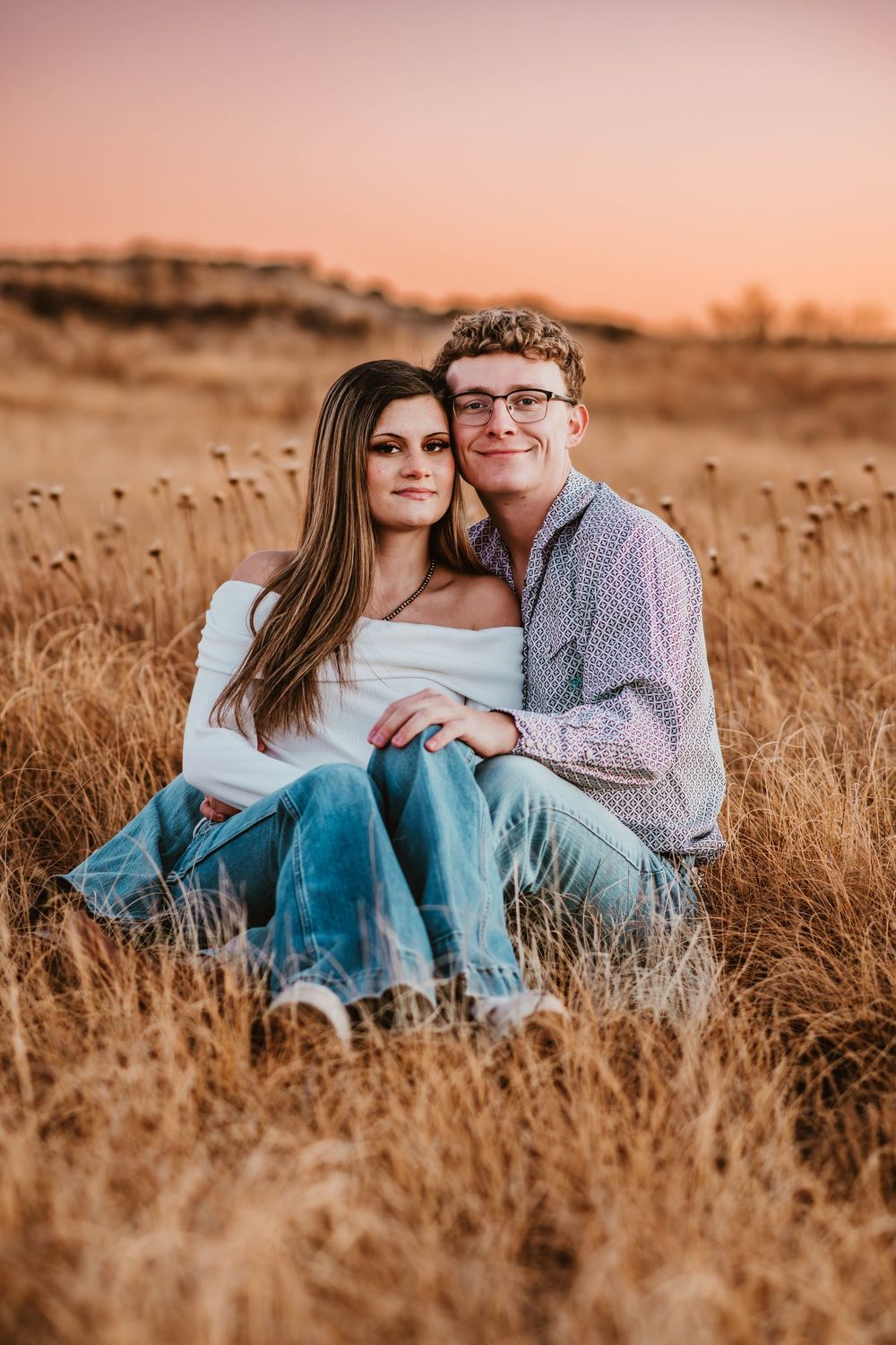 A romantic couple sits together in a golden wheat field during sunset wearing casual clothing.