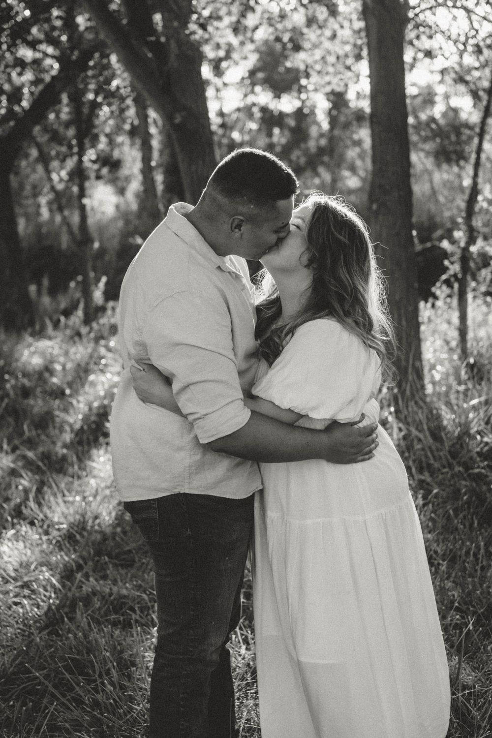 Black and white photo of a couple sharing a romantic kiss outdoors among trees, woman in white dress.