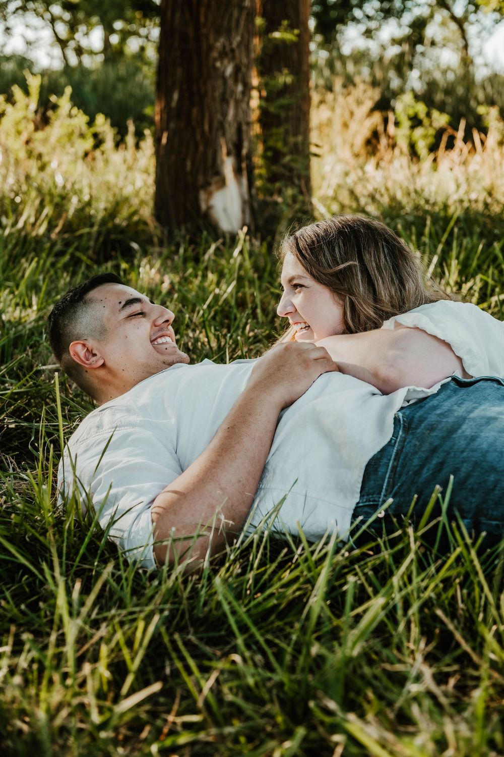 A couple laughs together while lying in green grass beneath a tree in golden sunlight.