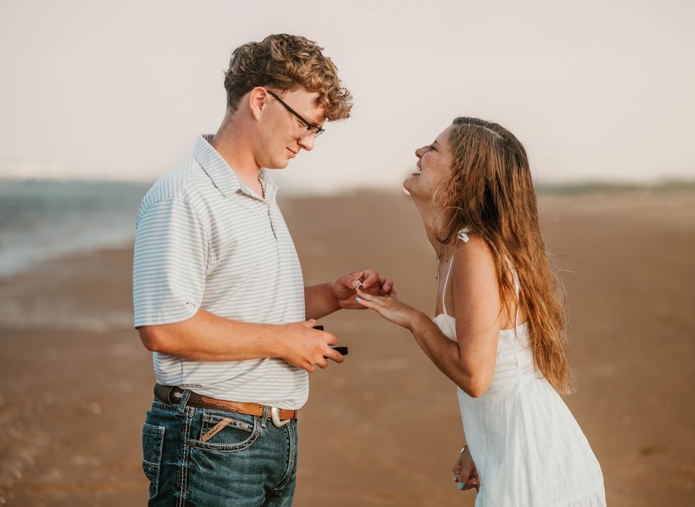 Man proposing to woman on a sandy beach at sunset, placing a ring on her finger as she laughs with joy.