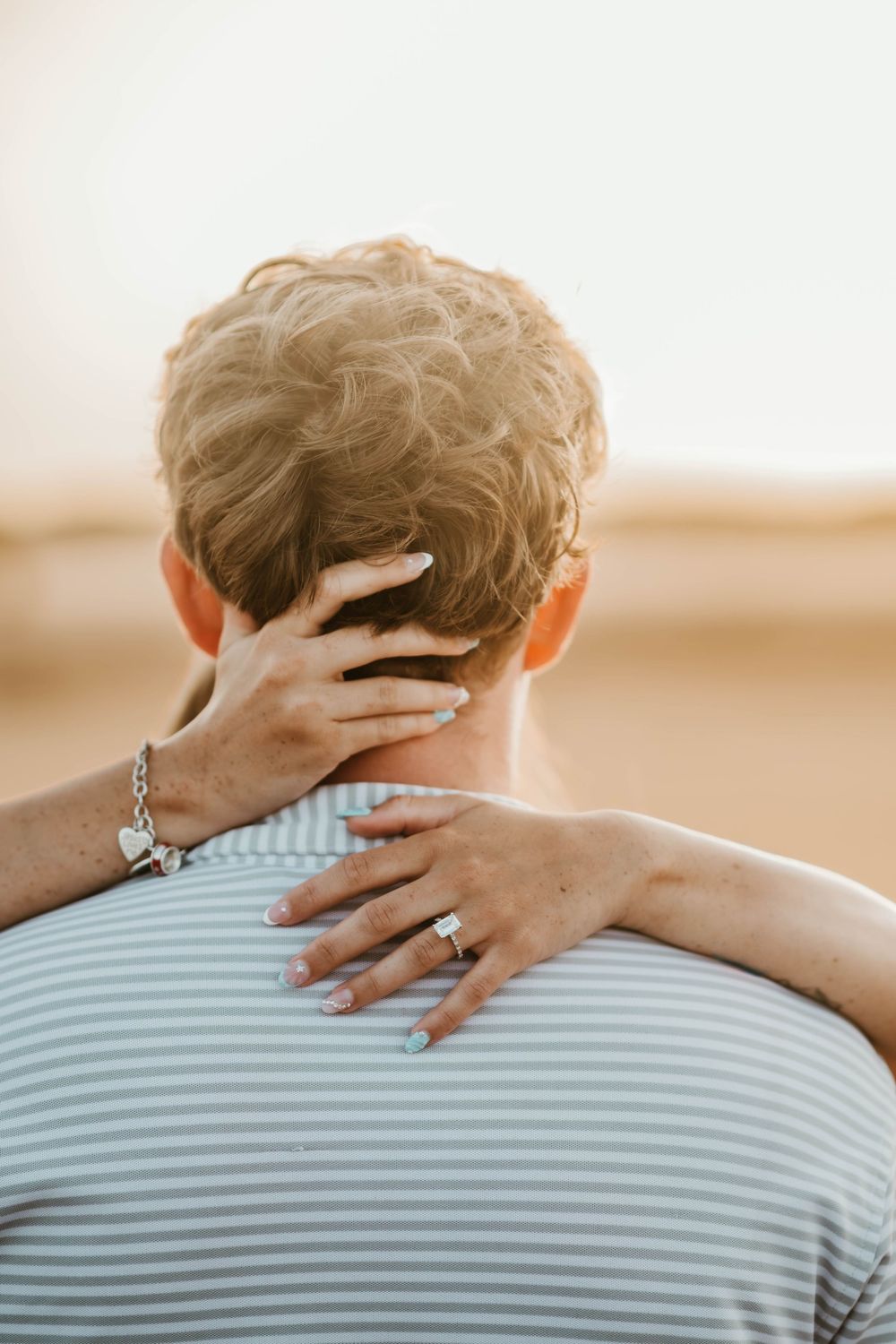 A woman with a bracelet and ring embraces someone from behind, resting her hand on their neck at sunset.