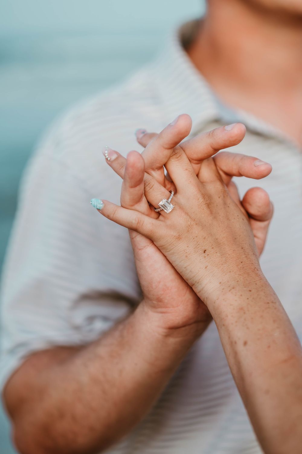 Close-up of couple holding hands with a diamond engagement ring on woman's finger with teal nail polish.