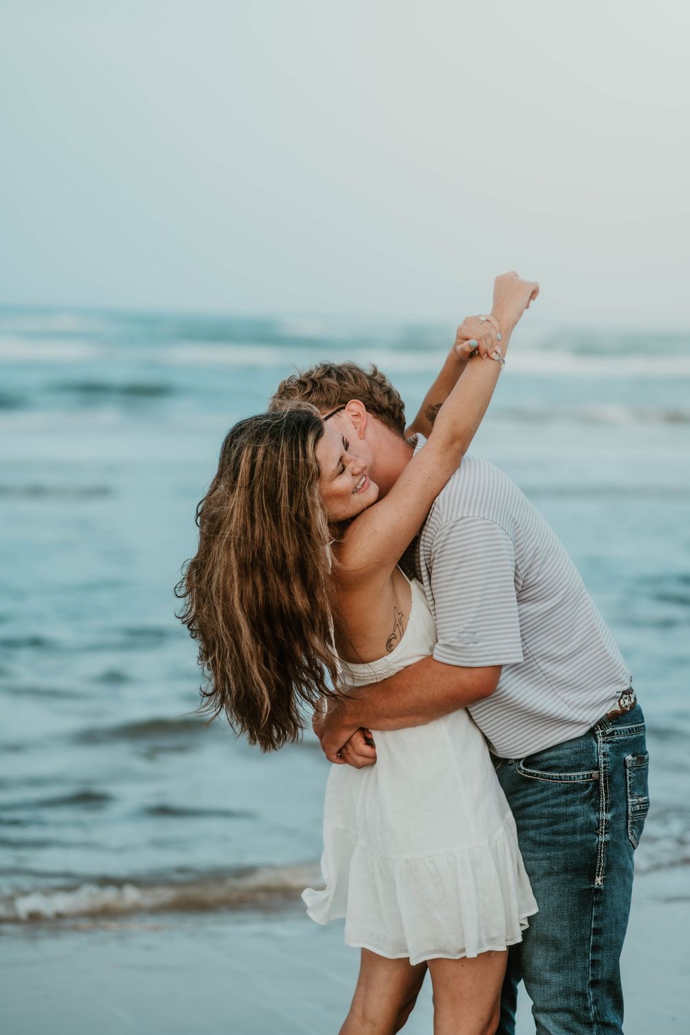 A romantic couple sharing a kiss on the beach, with the woman in a white dress and her arm raised joyfully.