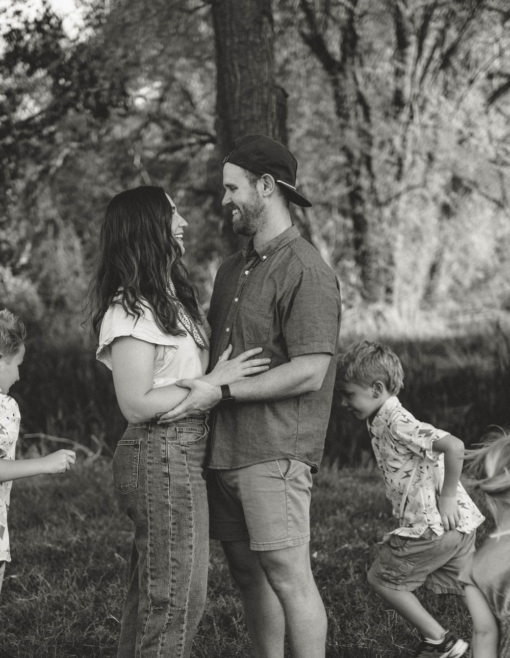 Black and white photo of a couple smiling at each other outdoors while children play around them near trees.