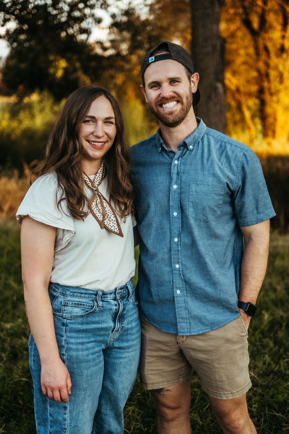 A smiling couple poses outdoors at golden hour, woman in white tee and jeans, man in blue shirt and khaki shorts.
