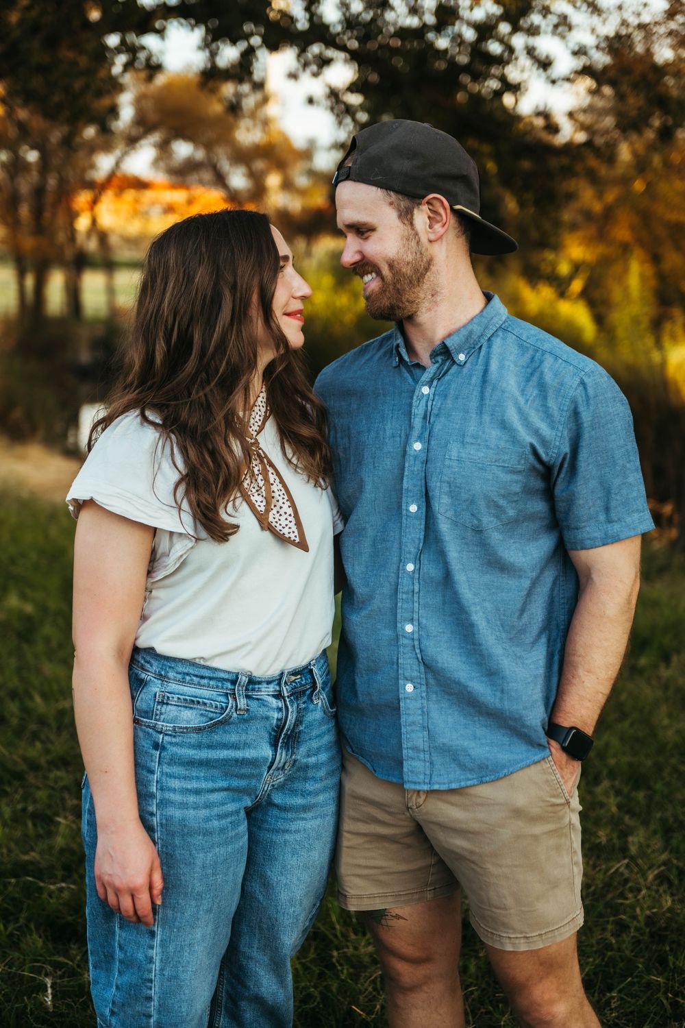 A smiling couple in casual attire gazes at each other outdoors during golden hour in a lush green park.