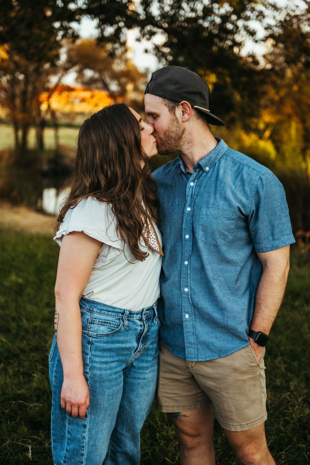 A couple sharing a kiss outdoors in a park during golden hour, woman in jeans and man in blue shirt and cap.