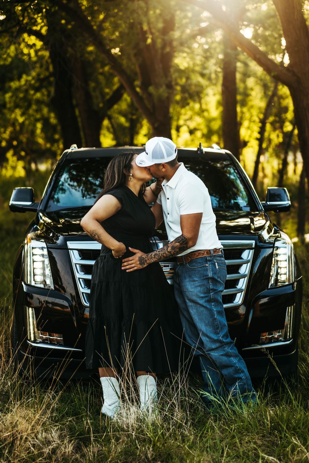 Couple sharing a romantic moment in front of a black Cadillac Escalade SUV in a sunlit forest setting.