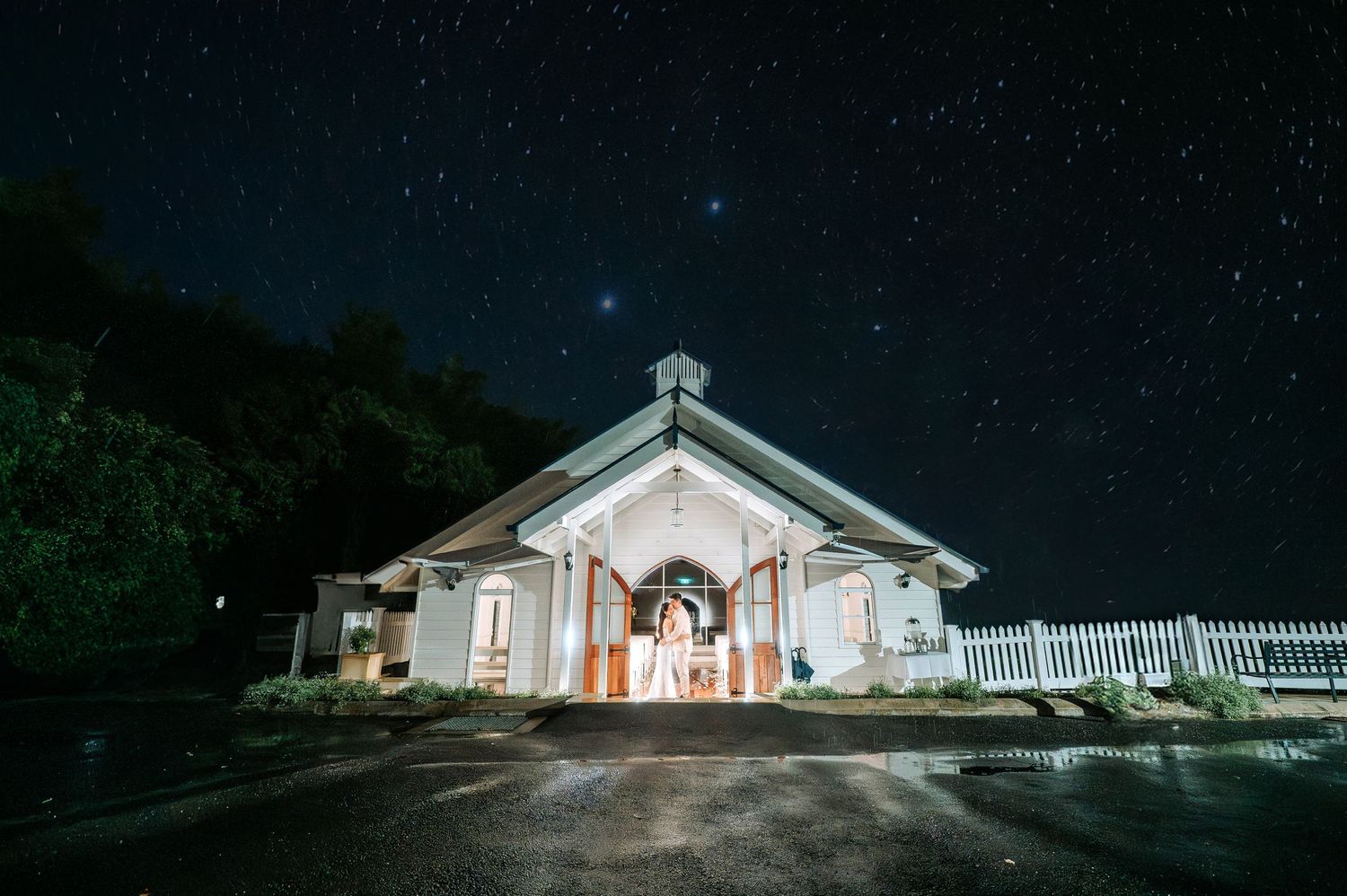 Small white chapel illuminated at night under a starry sky with warm interior lights glowing through entrance doors.