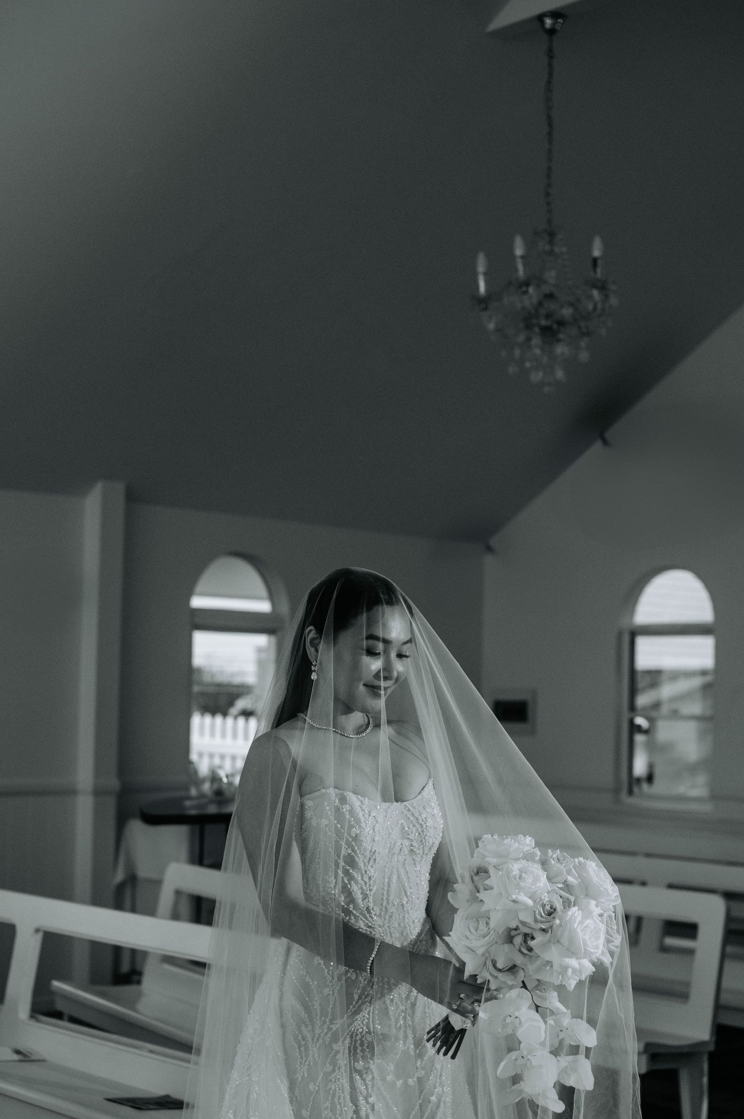 A bride in a delicate white lace gown holds a bouquet while standing in a chapel with arched windows.