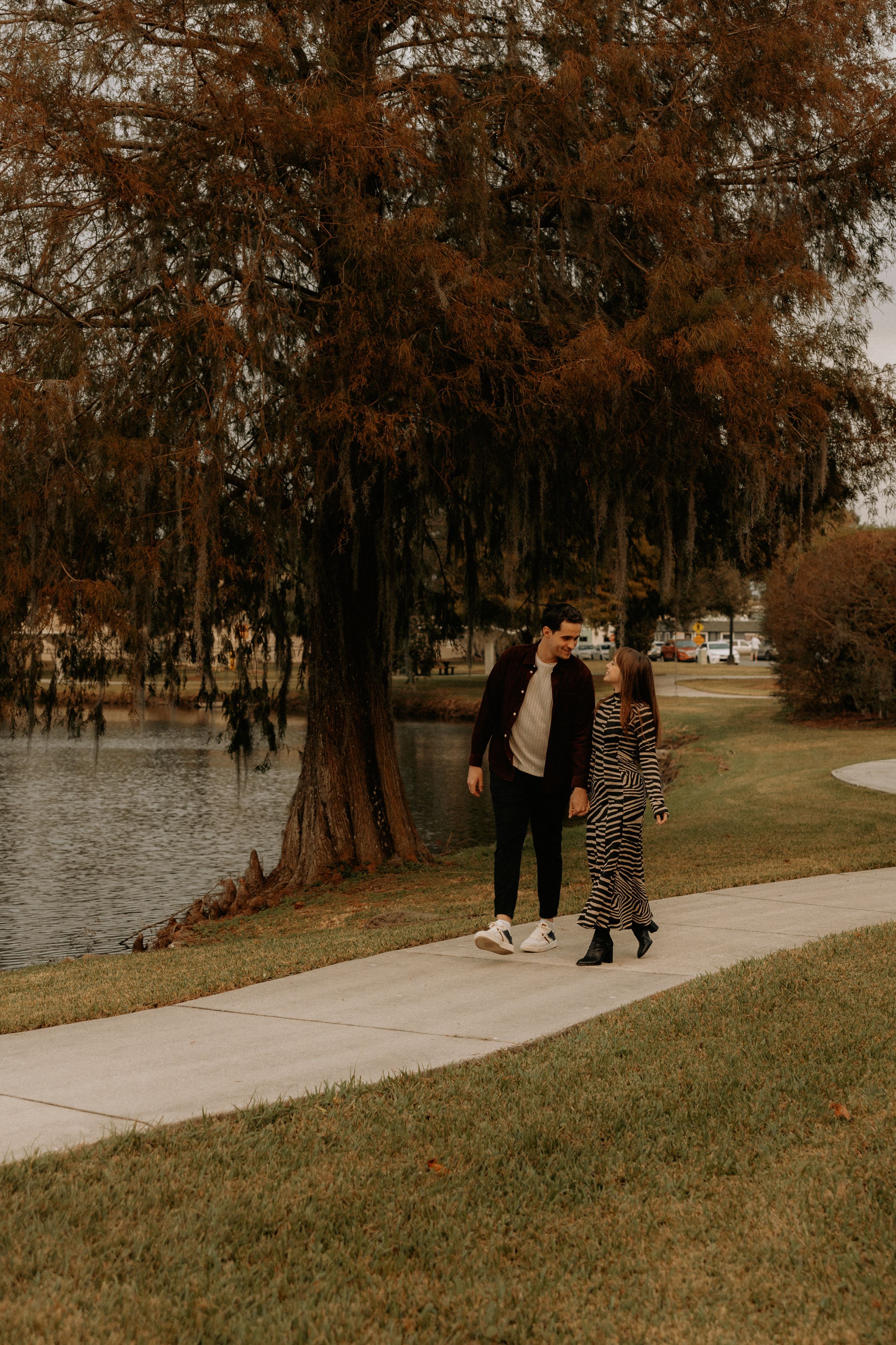 Two people stroll together on a concrete pathway beside a lake with fall foliage.