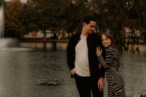 A couple in coordinated outfits walks along a lakeside path during autumn.