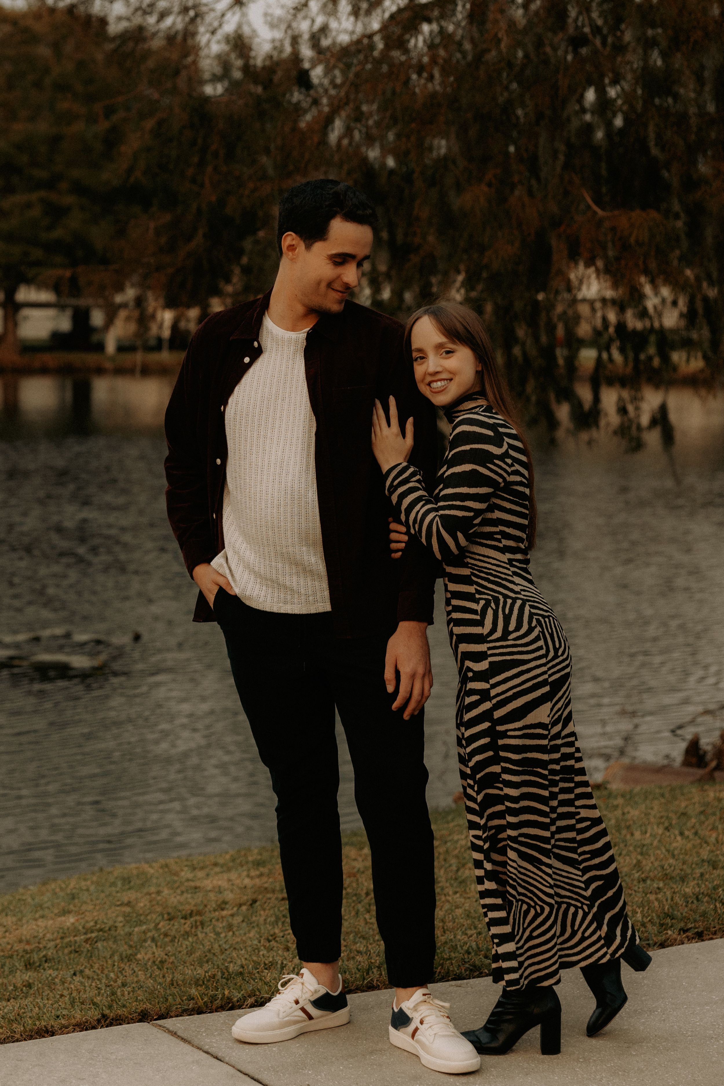 A couple shares a romantic moment on a waterfront walkway with autumn trees in the background.
