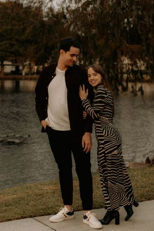 A couple shares a romantic moment on a waterfront walkway with autumn trees in the background.