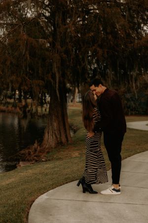 A couple poses together on a concrete path with a lake and trees in the background.