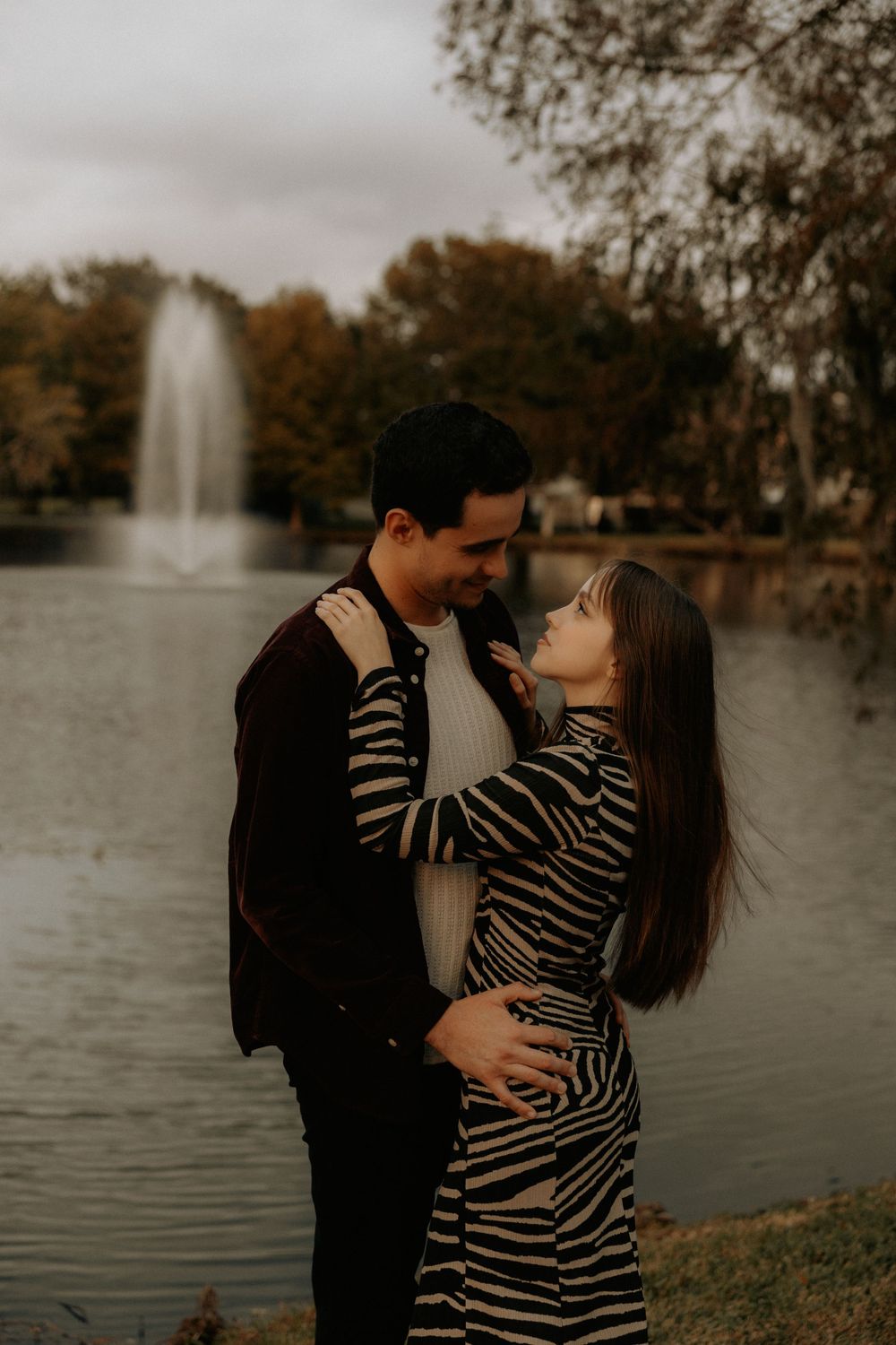 Romantic couple embracing by a lake with a fountain in the background during sunset.