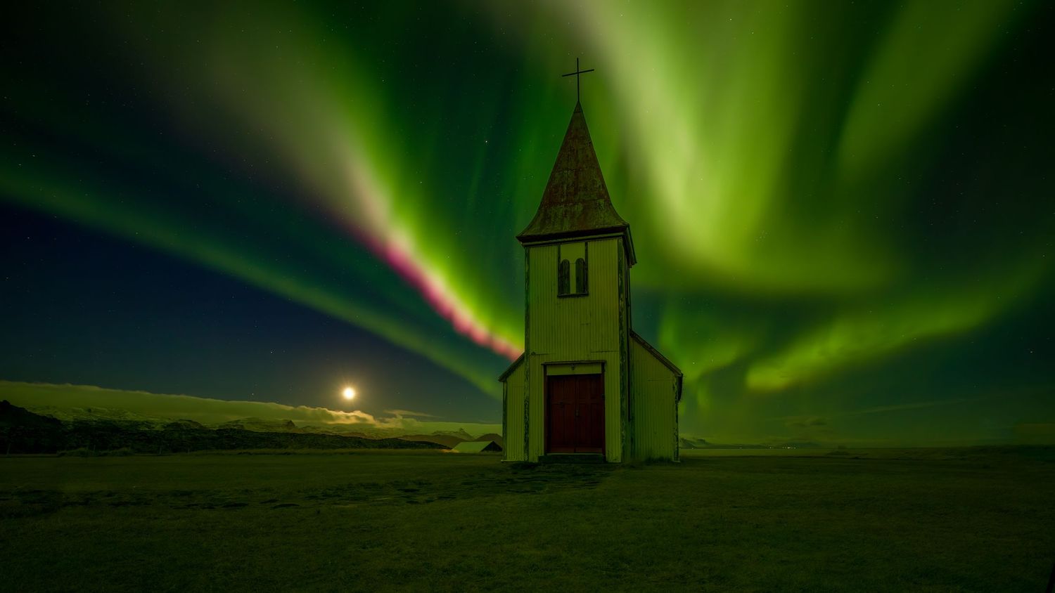 The Northern lights dance across the sky overhead and cast a green hue to a church and Jupiter in the background in Iceland