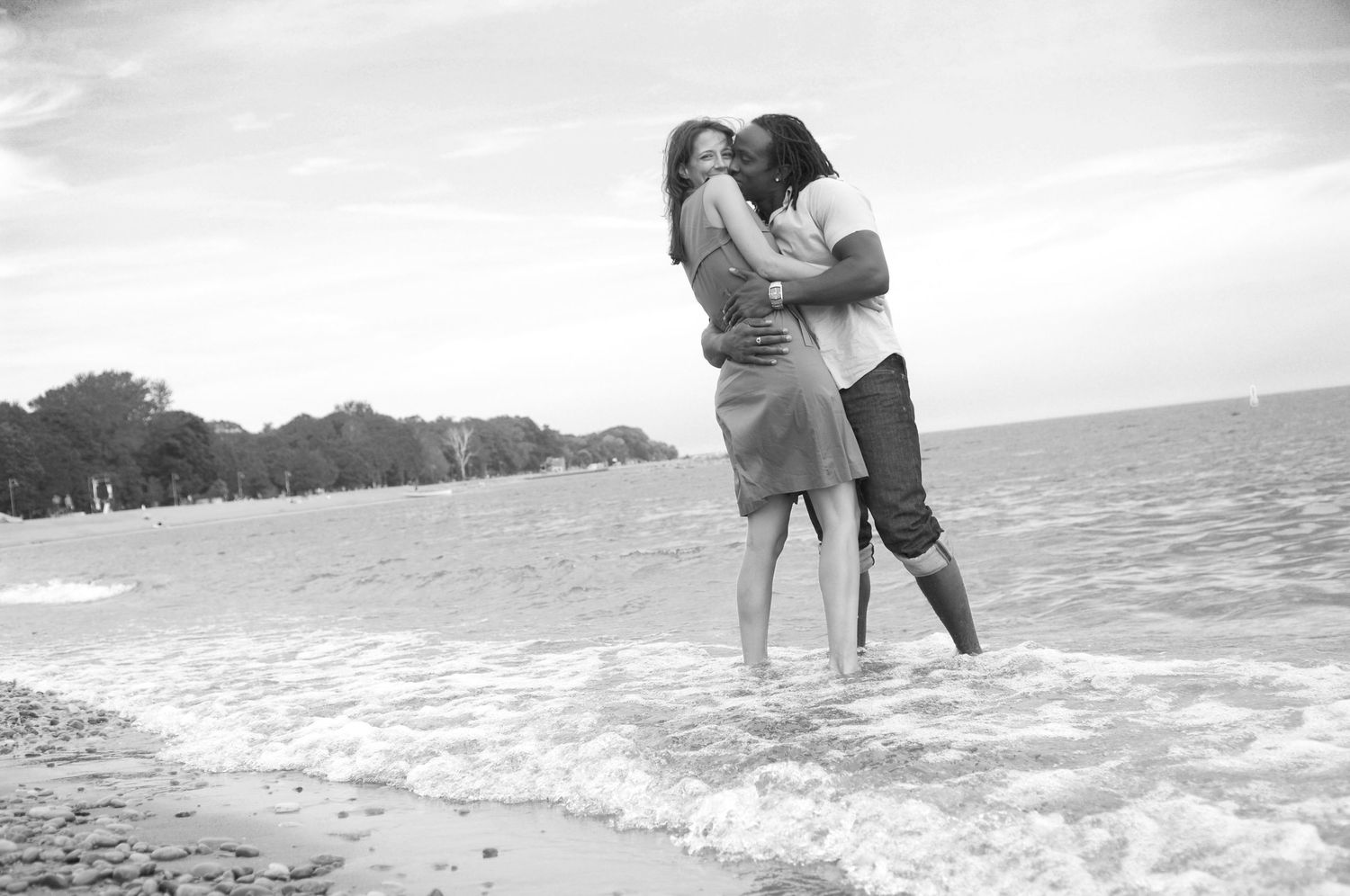 A romantic couple embraces on a beach as waves lap at their feet.
