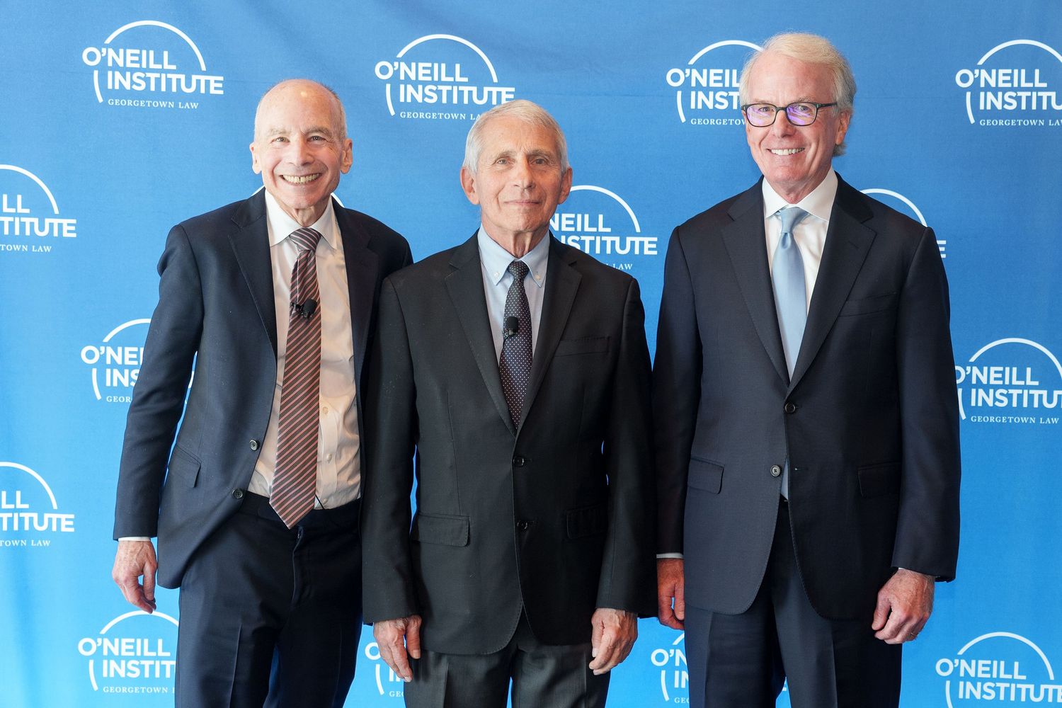 Three people in business suits pose together at a blue-branded O'Neill Institute event backdrop.
